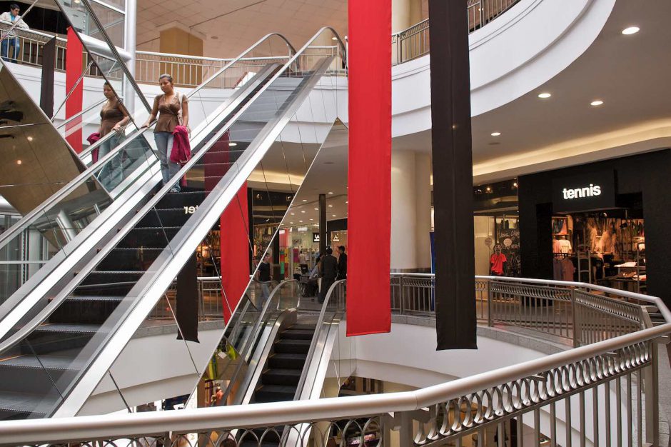 Kleurfoto van 2 in kostuum geklede mannen die de straat oversteken ter hoogte van shopping center El Jardin