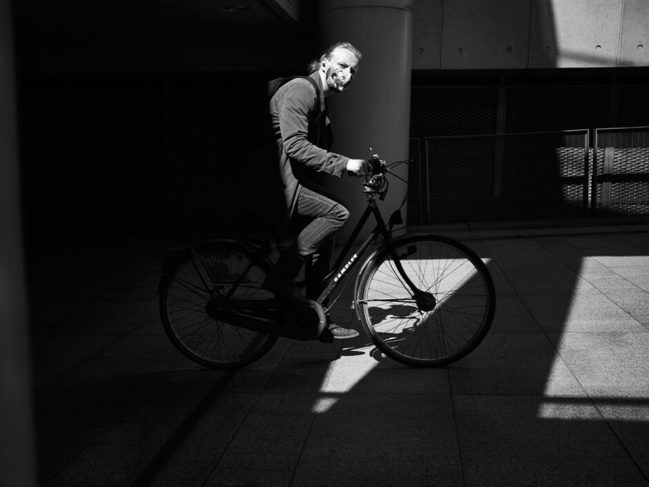 Black & white street photography of homo urbanus, more precisely of a young man on a bike entering the image in a beam of light