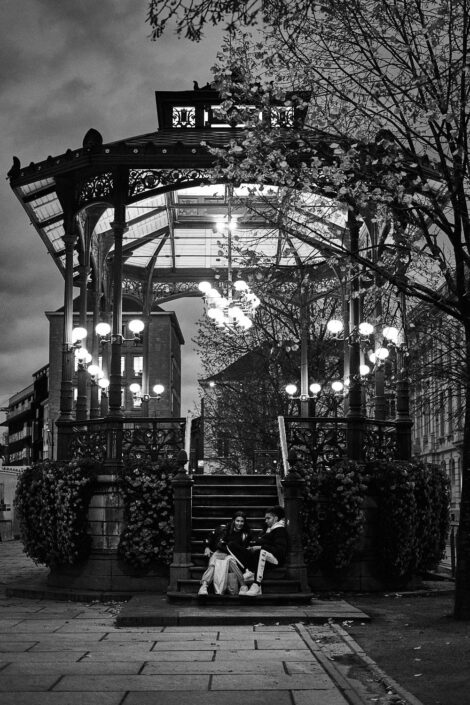 Black & white street photography of homo urbanus, more precisely of two youngsters at the foot of a music kiosk at dusk.