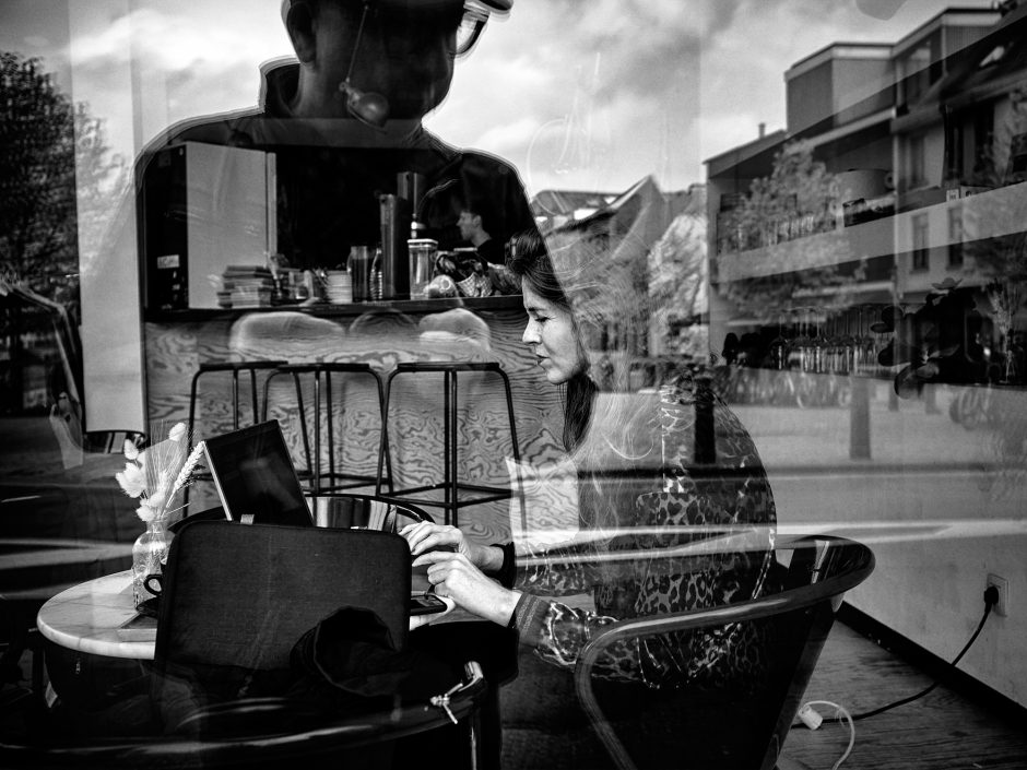 Black & white photograph of a young woman working on her laptop in a coffee shop while the reflection of the photographer is looming over her