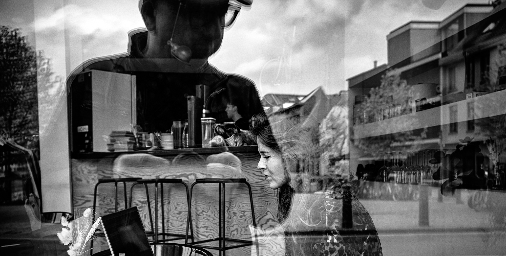 Black & white photograph of a young woman working on her laptop in a coffee shop while the reflection of the photographer is looming over her