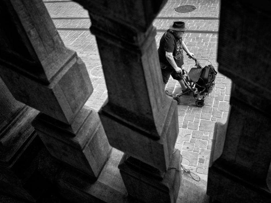 Black & white photograph of a elderly man with a hat and a rock 'n roll t-shirt, pushing a rollator